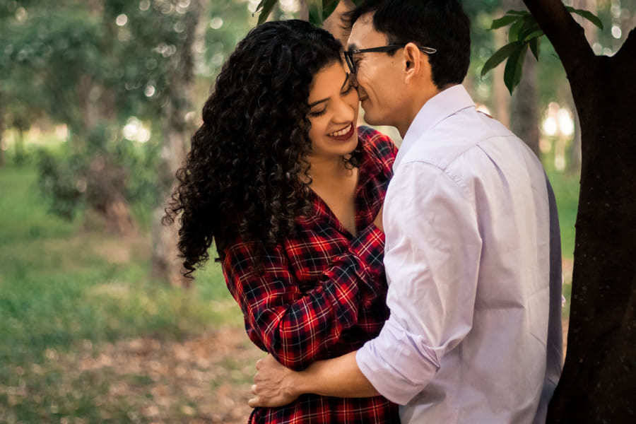 ENSAIO FOTOGRAFICO PRE CASAMENTO OU PRE WEDDING AO AR LIVRE EM BOSQUE DAS GREVILEAS GREVILIAS MARINGA NO PARANA NA IMAGEM O NOIVO ESTA ENCONTADO EM UMA ARVORE DANDO UM CHEIRINHO NO CABELO DA NOIVA QUE ESTA SORRINDO