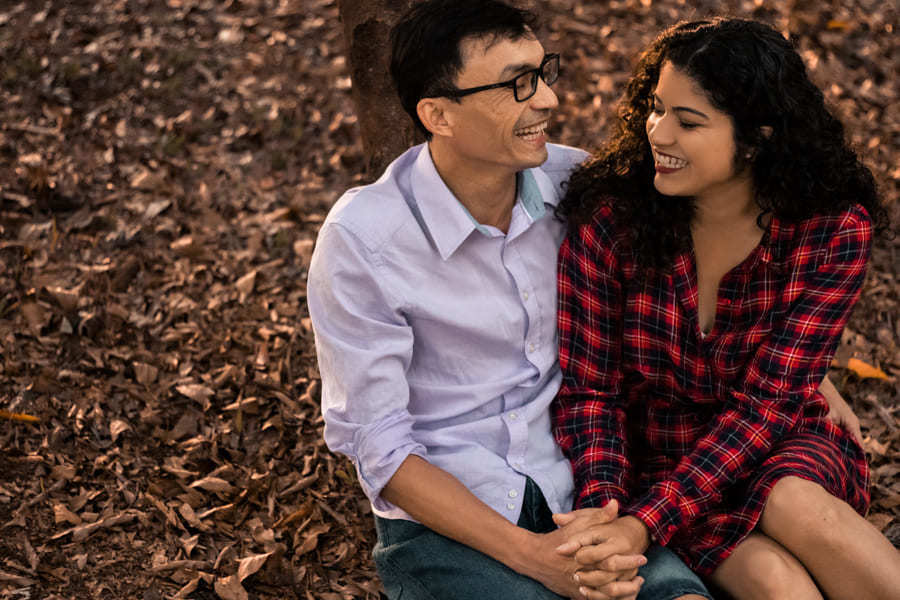 ENSAIO FOTOGRAFICO PRE CASAMENTO OU PRE WEDDING AO AR LIVRE EM BOSQUE DAS GREVILEAS GREVILIAS MARINGA NO PARANA NA IMAGEM O CASAL ESTA SENTADO LADO A LADO EM MEIO A VEGETACAO MARROM COM CLIMA DE OUTONO AMBOS SORRINDO