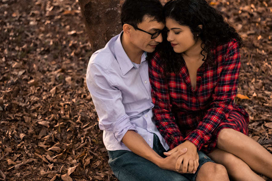 ENSAIO FOTOGRAFICO PRE CASAMENTO OU PRE WEDDING AO AR LIVRE EM BOSQUE DAS GREVILEAS GREVILIAS MARINGA NO PARANA NA IMAGEM O CASAL ESTA SENTADO LADO A LADO EM MEIO A VEGETACAO MARROM COM CLIMA DE OUTONO DE CABEÇAS ENCOSTADAS E DE OLHOS FECHADOS EM ORACAO