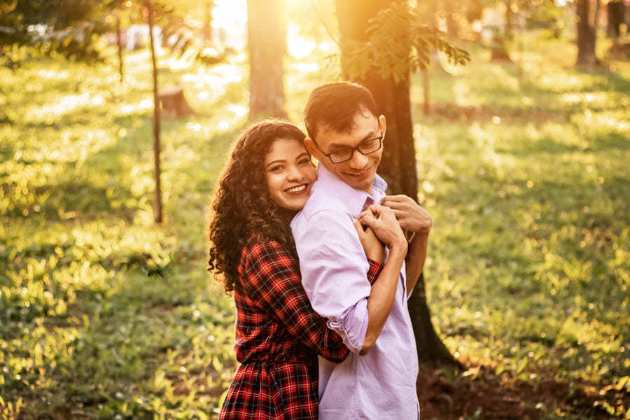 ENSAIO FOTOGRAFICO PRE CASAMENTO OU PRE WEDDING AO AR LIVRE EM BOSQUE DAS GREVILEAS GREVILIAS MARINGA NO PARANA NA IMAGEM A NOIVA ABRACA O NOIVO POR TRAS SEGURANDO O SEU PEITO E OLHANDO PARA A CAMERA