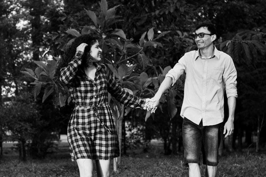 ENSAIO FOTOGRAFICO PRE CASAMENTO OU PRE WEDDING AO AR LIVRE EM BOSQUE DAS GREVILEAS GREVILIAS MARINGA NO PARANA NA IMAGEM O CASAL ESTA CAMINHANDO DE MAOS DADAS ENQUANTO O NOIVO OLHA PARA A NOIVA ELA RETRIBUI E SEGURA O CABELO FOTO EM PRETO E BRANCO