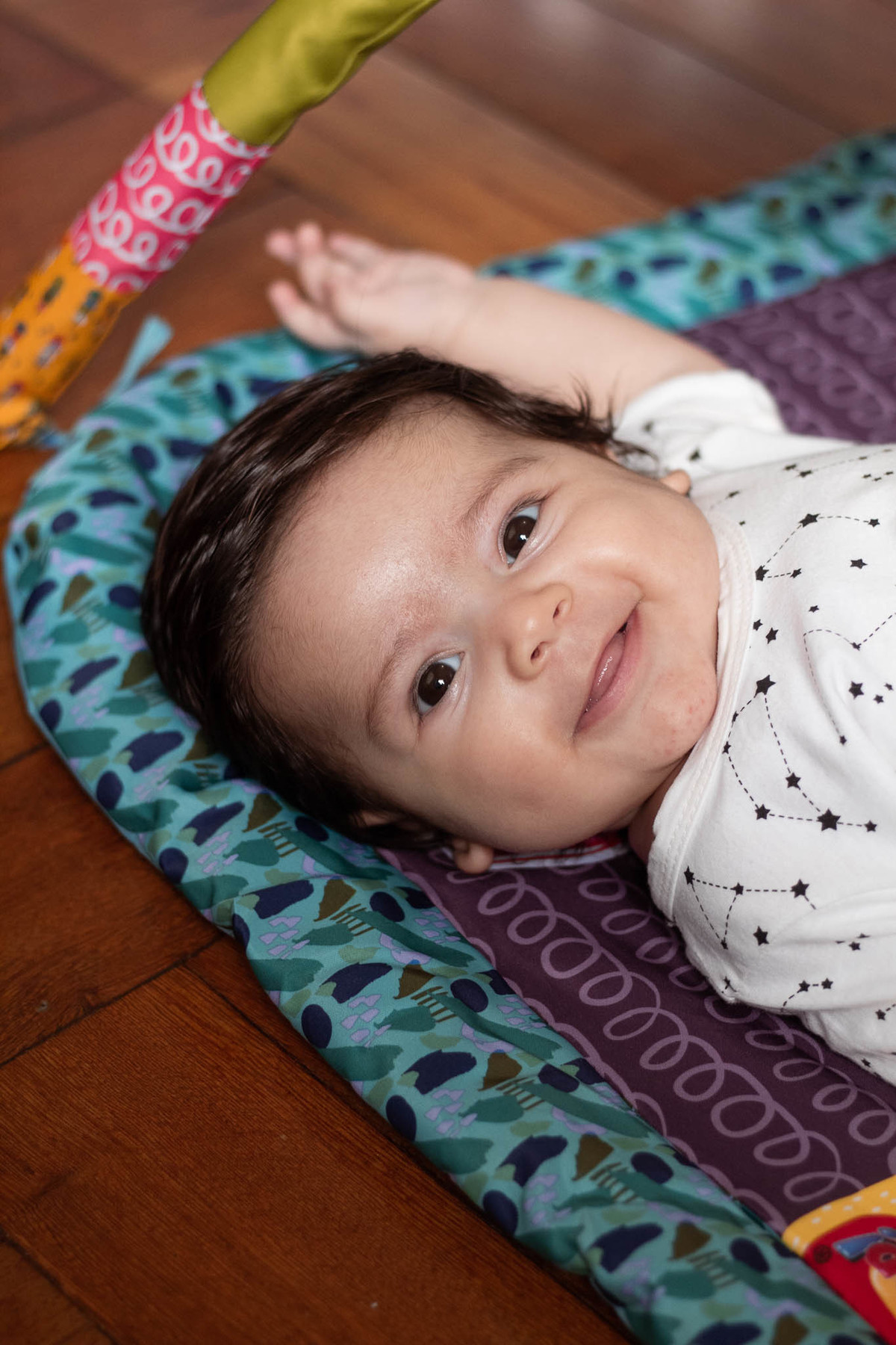 bebê de pele clara, cabelos e olhos castanho escuro, sorrindo, deitado num tapete colorido.