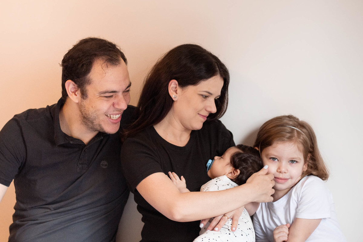 Família de quatro pessoas: pai, mãe, menina de 5 anos e bebê de 3 meses. O pai e a mãe olham para a família sorrindo, o bebê dorme e a criança olha para a câmera sorrindo.