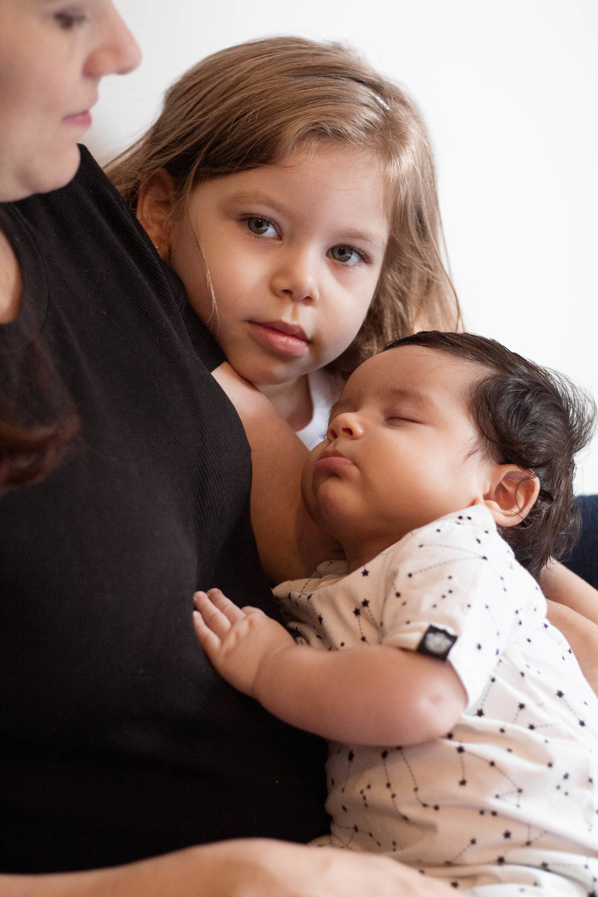 Foto de um bebê de pele clara, cabelos e olhos castanho escuro, dormindo nos braços da mãe, com menina de 5 anos ao fundo olhando para a câmera.