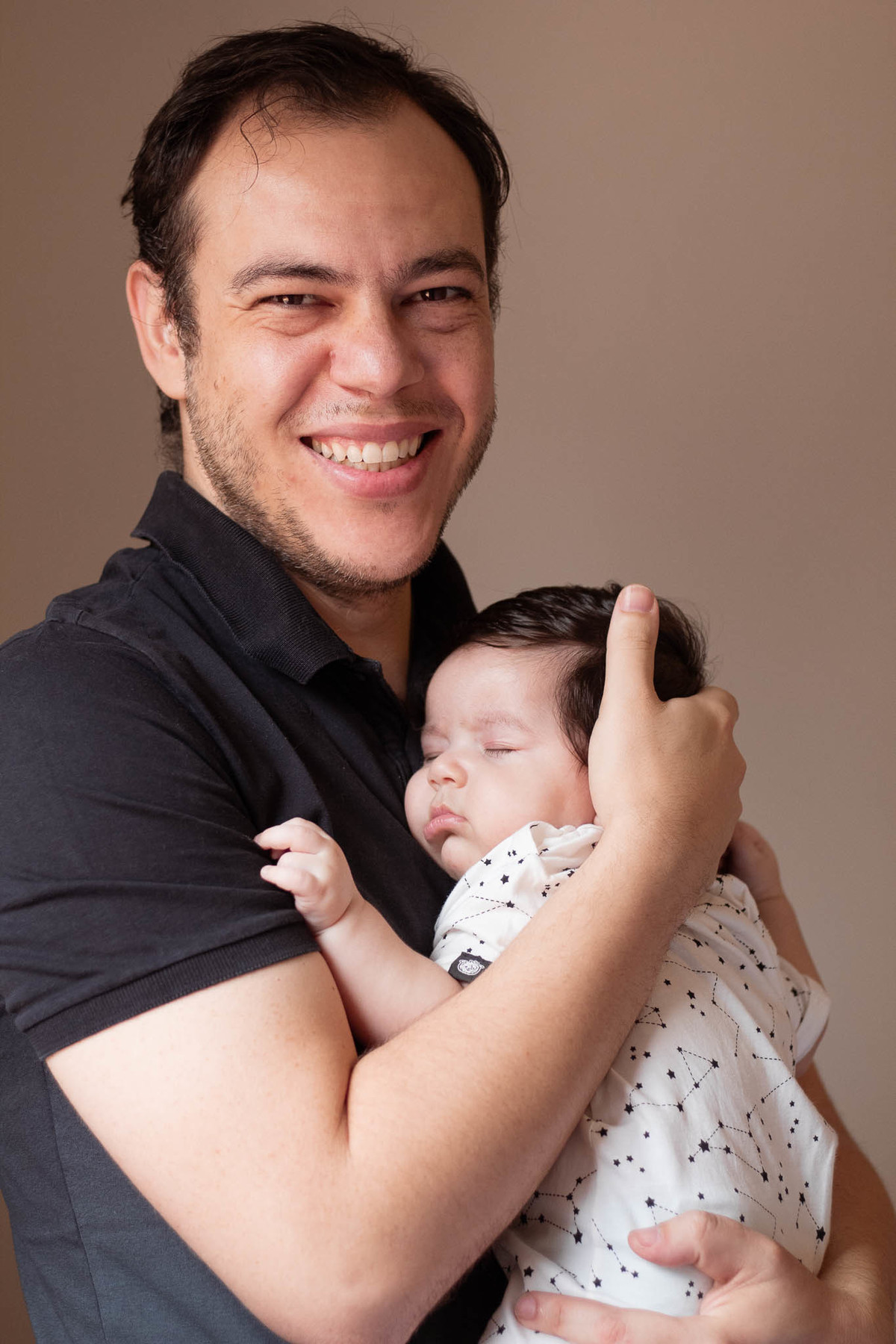 Pai sorrindo olhando para a câmera segurando seu bebê de pele clara, cabelos e olhos castanho escuro, enquanto dorme.