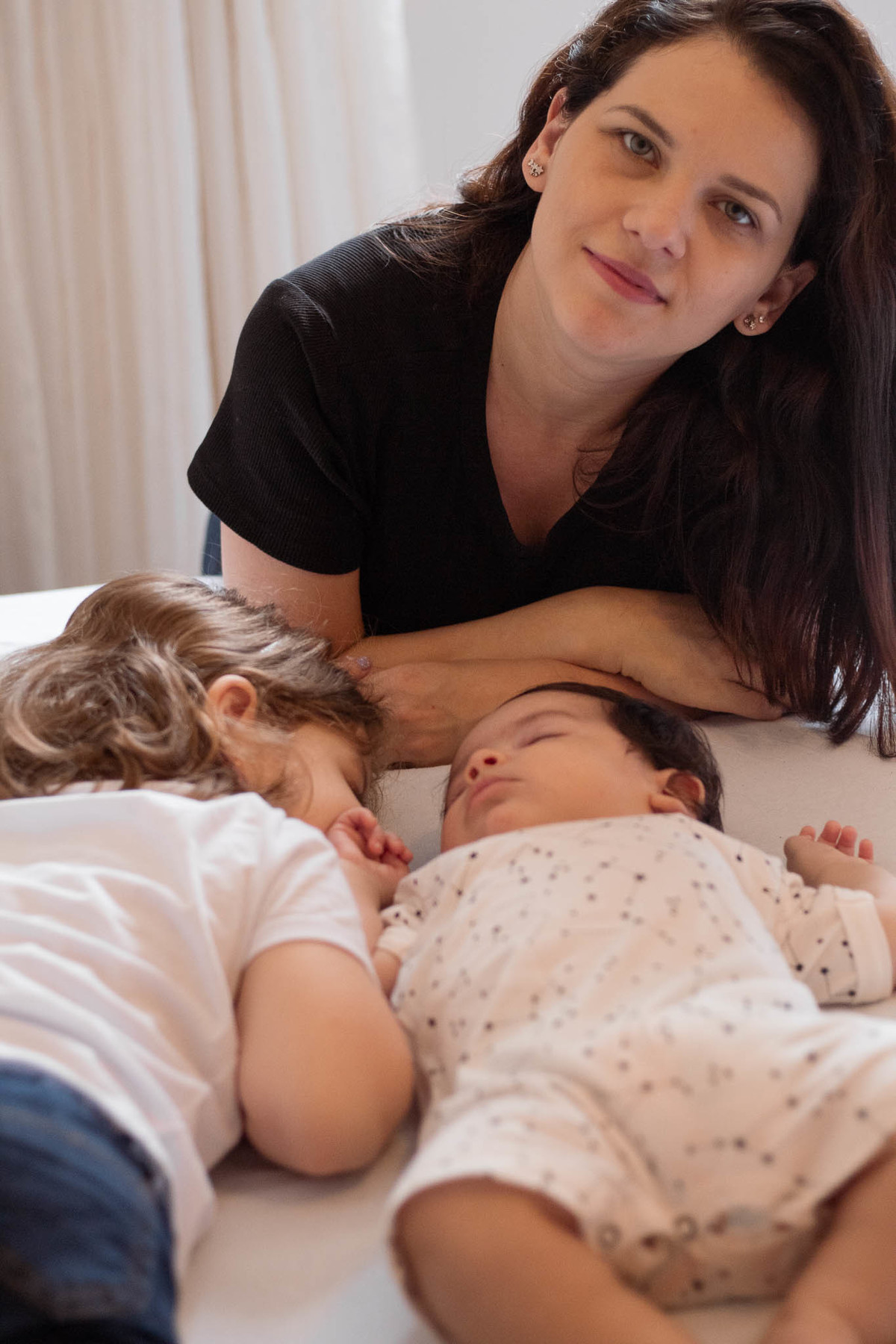 bebê de pele clara, cabelos e olhos castanho escuro, dormindo com sua irmã, uma menina loira de 5 anos, enquanto sua mãe olha para a câmera.