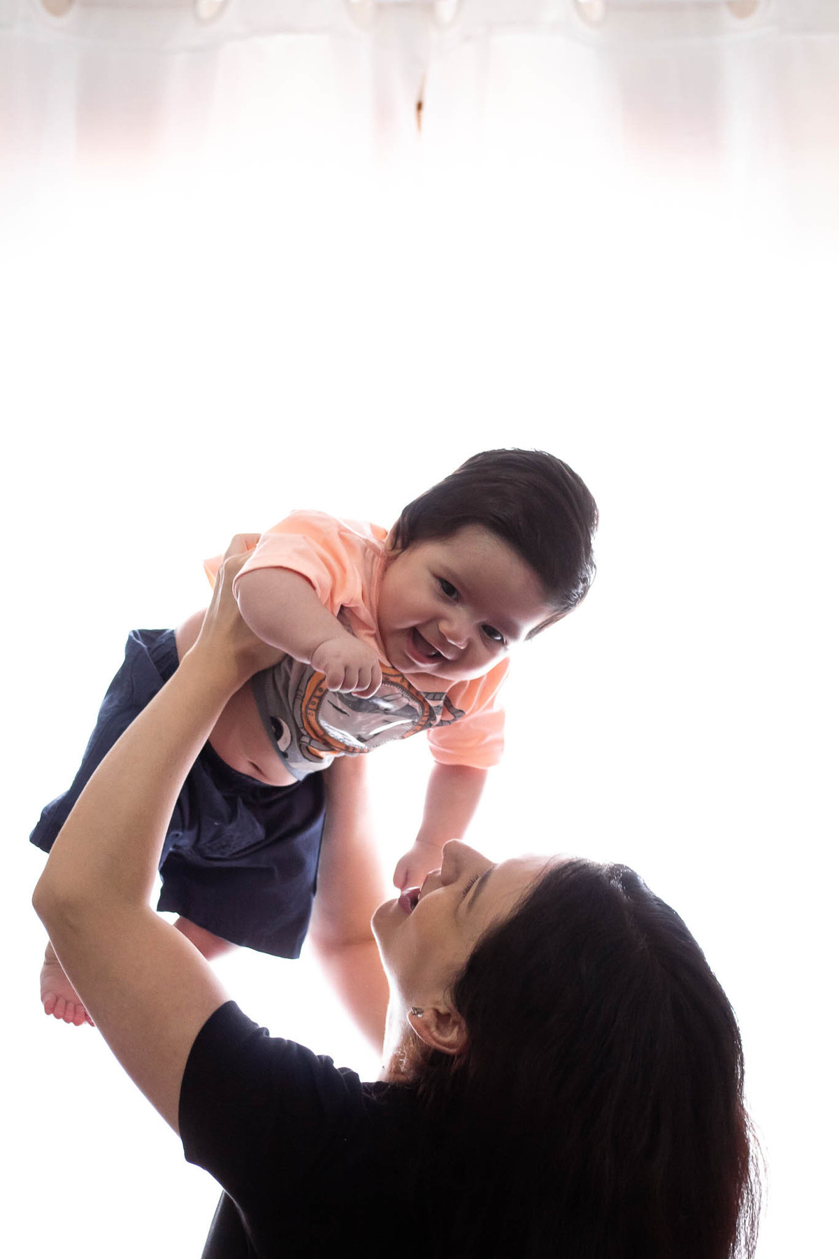 bebê de pele clara, cabelos e olhos castanho escuro, sendo levantado pela mamãe, ambos estão sorrindo.
