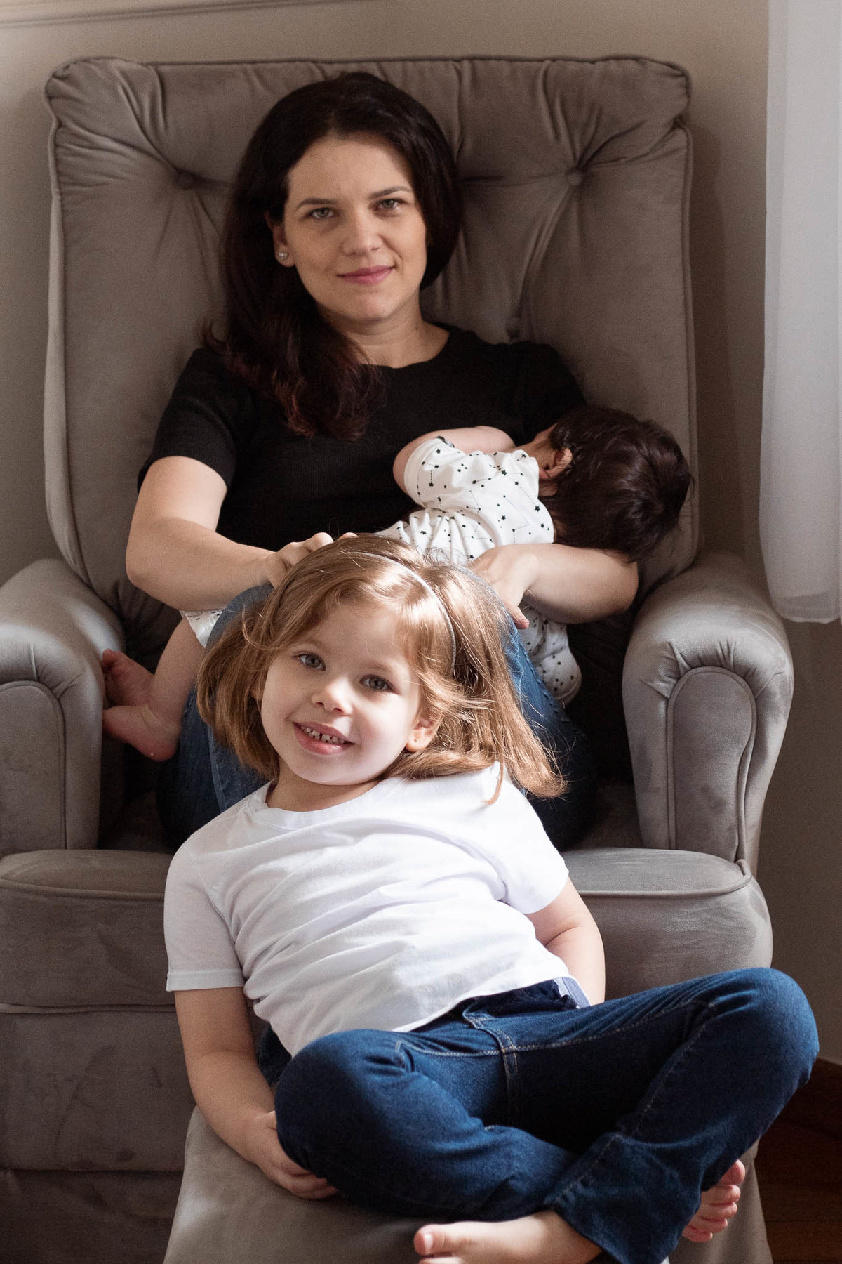 Foto em família. Mãe amamentando o bebê sentada em uma poltrona cinza e menina de 5 anos, loira, sorrindo, olhado para a câmera, sentada na frente da mãe.