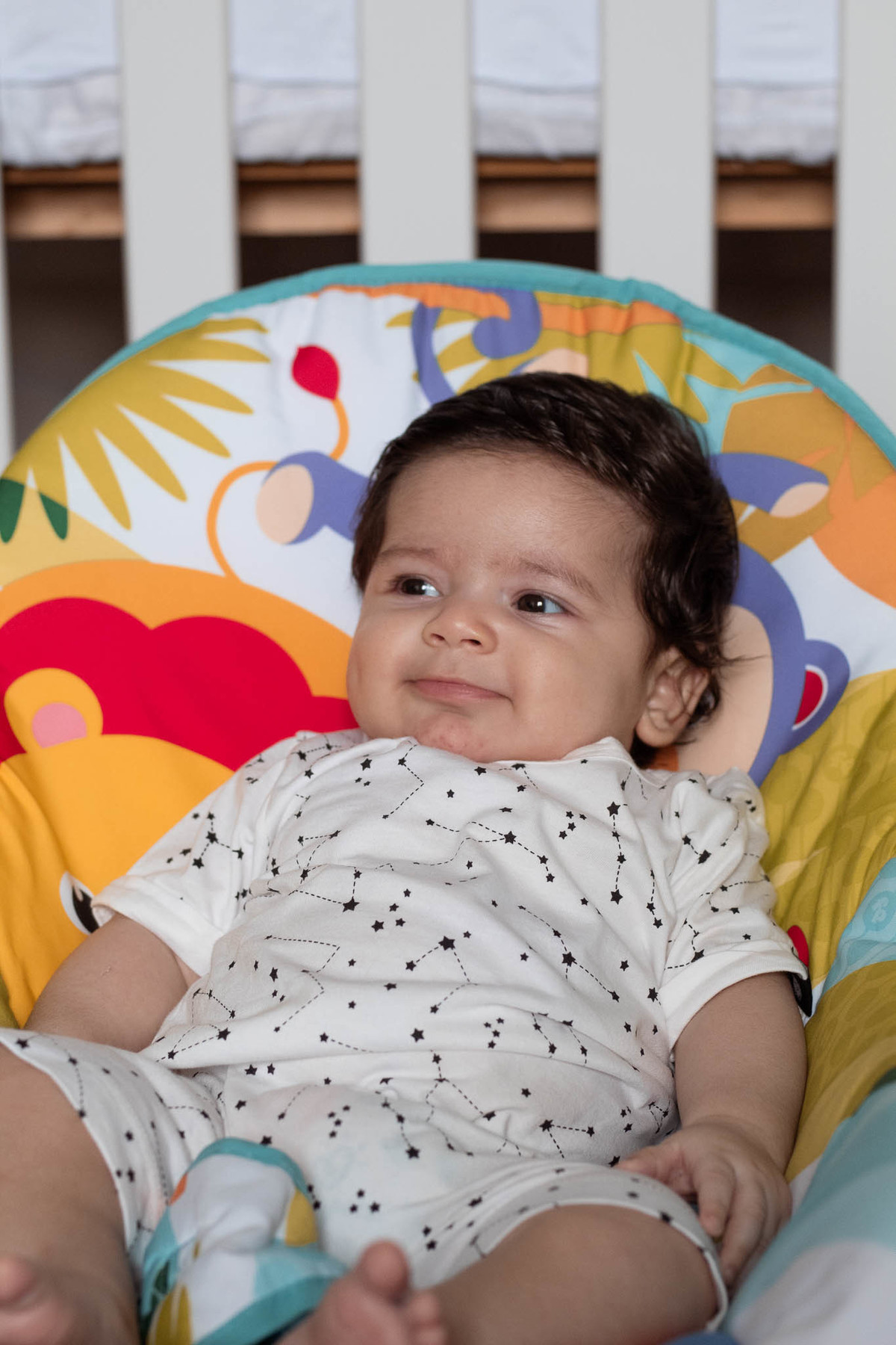 bebê de pele clara, cabelos e olhos castanho escuro, sentado numa cadeirinha colorida olhando para o lado.