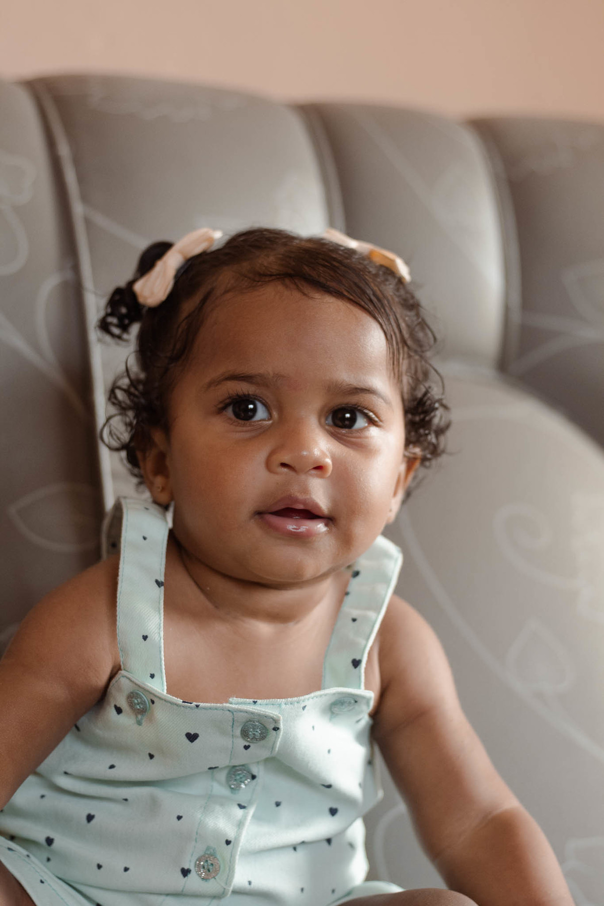 bebê de 1 ano, menina, pele parda, cabelo cacheado castanho e olhos castanhos, vestindo macacão azul e lacinho rosa na cabeça, olhando para a câmera