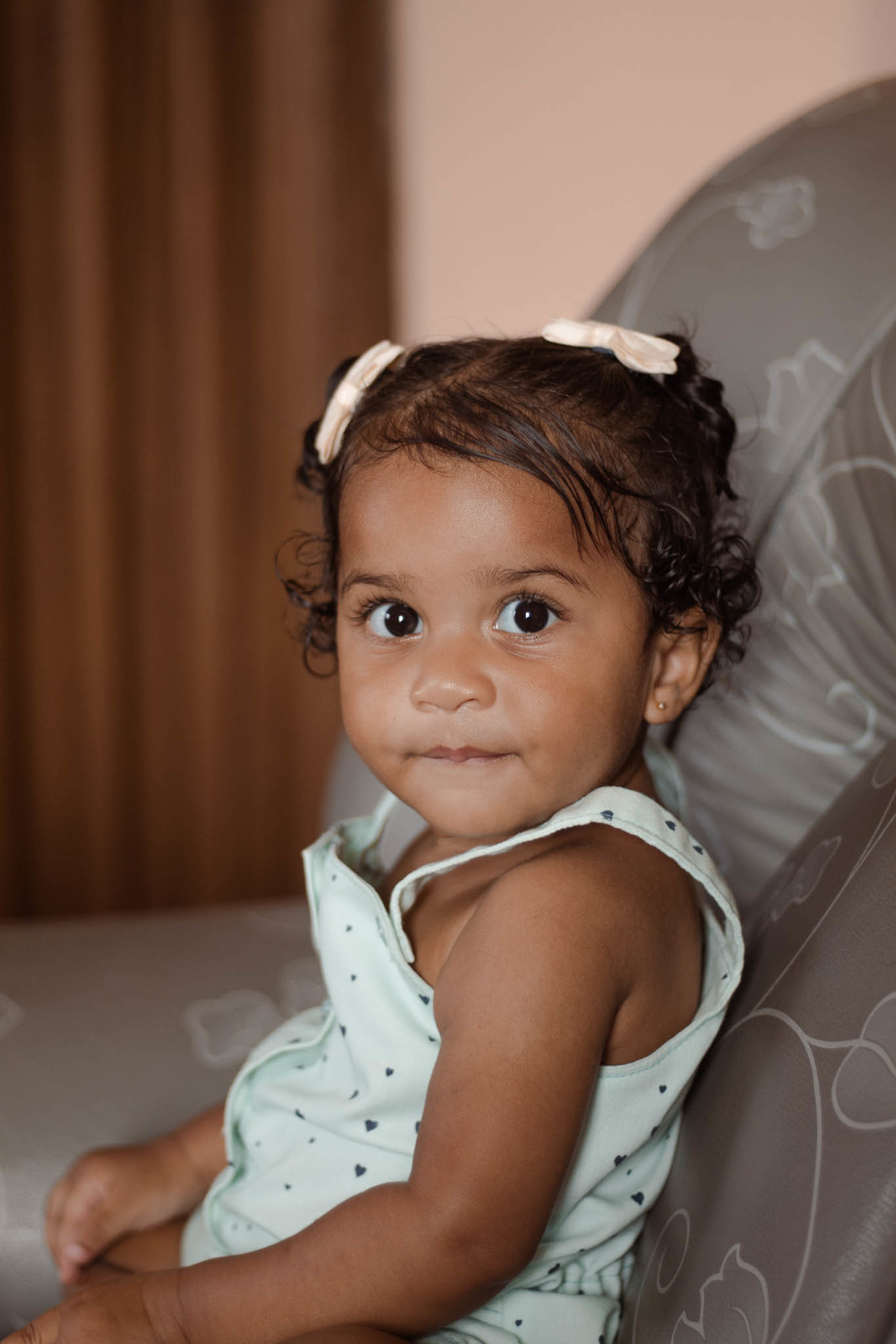bebê de 1 ano, menina, pele parda, cabelo cacheado castanho e olhos castanhos, vestindo macacão azul e lacinho rosa na cabeça, olhando para a câmera