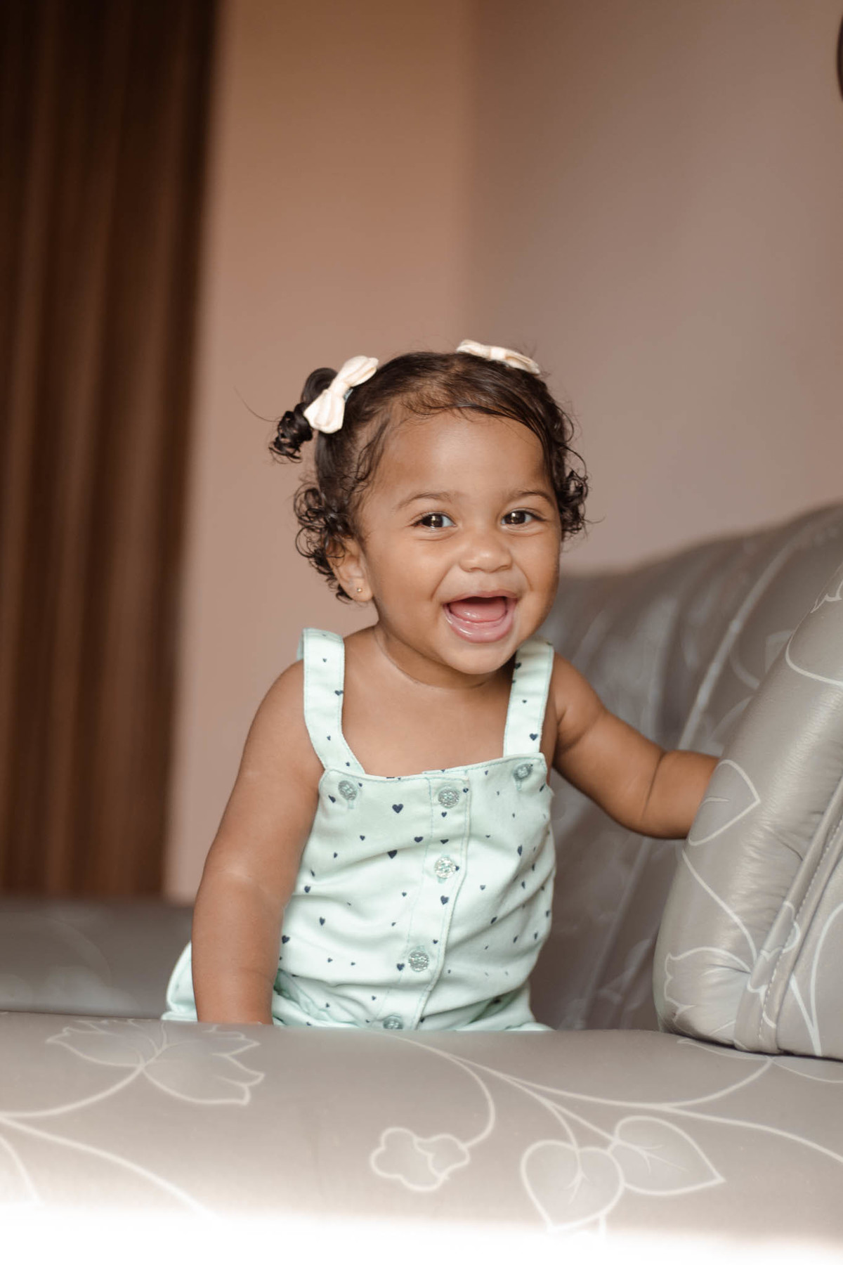 bebê de 1 ano, menina, pele parda, cabelo cacheado castanho e olhos castanhos, vestindo macacão azul e lacinho rosa na cabeça, sorrindo apoiada em um sofá cinza