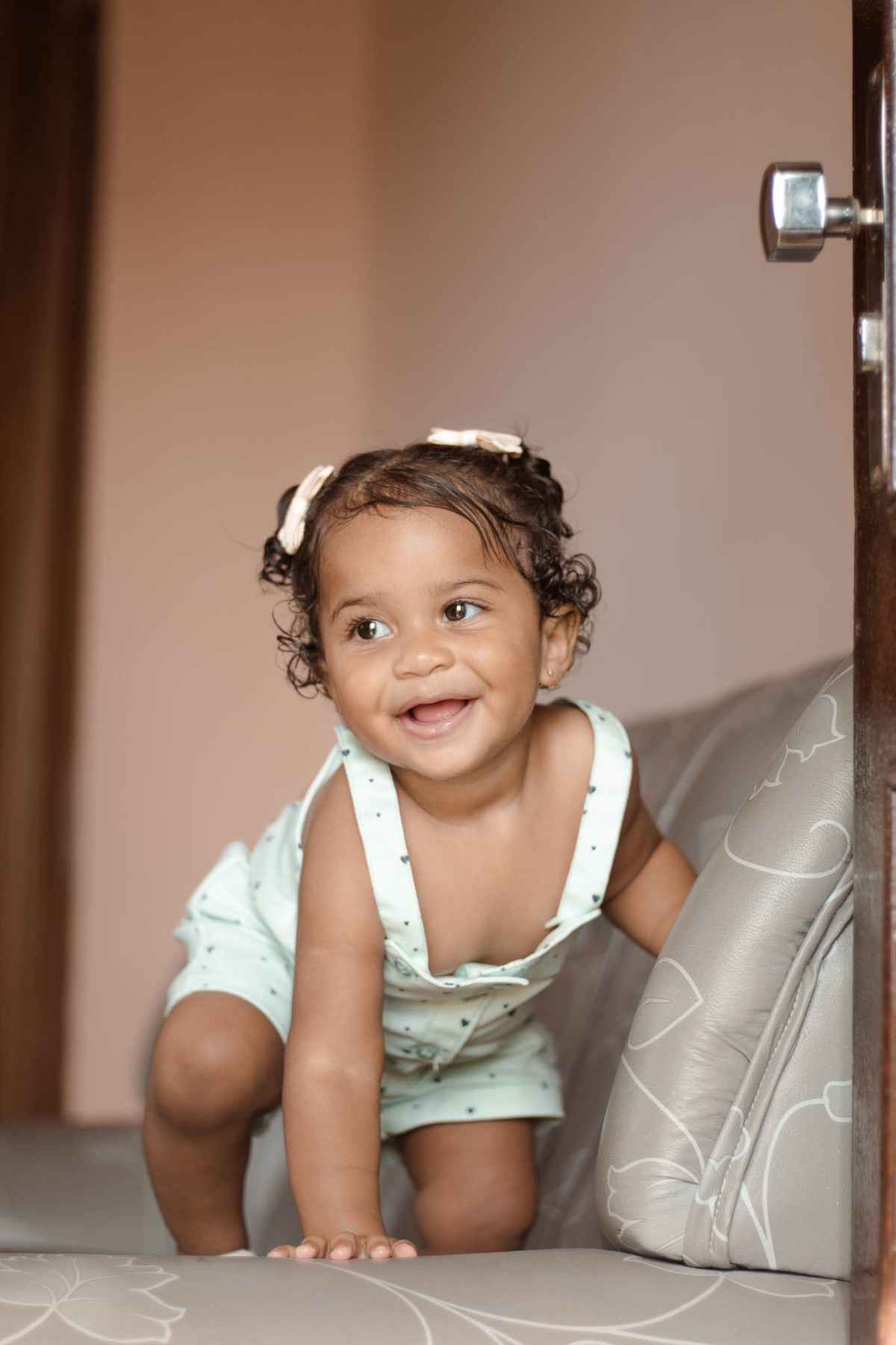 bebê de 1 ano, menina, pele parda, cabelo cacheado castanho e olhos castanhos, vestindo macacão azul e lacinho rosa na cabeça sorrindo olhando para o lado