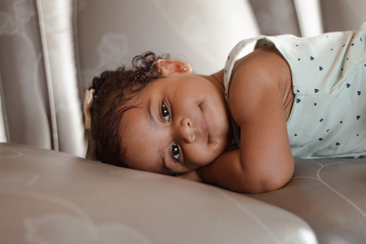 bebê de 1 ano, menina, pele parda, cabelo cacheado castanho e olhos castanhos, vestindo macacão azul e lacinho rosa na cabeça, deitada em um sofá cinza olhando para a câmera 