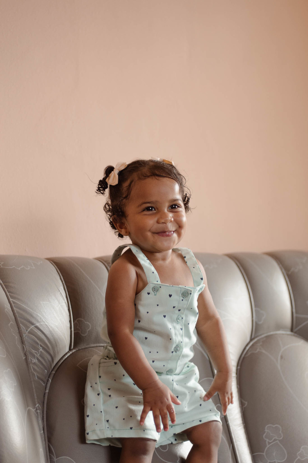bebê de 1 ano, menina, pele parda, cabelo cacheado castanho e olhos castanhos, vestindo macacão azul e lacinho rosa na cabeça, em pé em um sofá cinza e sorrindo olhado para a frente