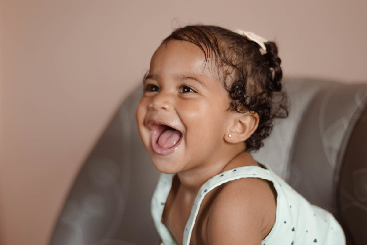 bebê de 1 ano, menina, pele parda, cabelo cacheado castanho e olhos castanhos, vestindo macacão azul e lacinho rosa na cabeça, em pé em um sofá cinza e sorrindo olhado para o lado