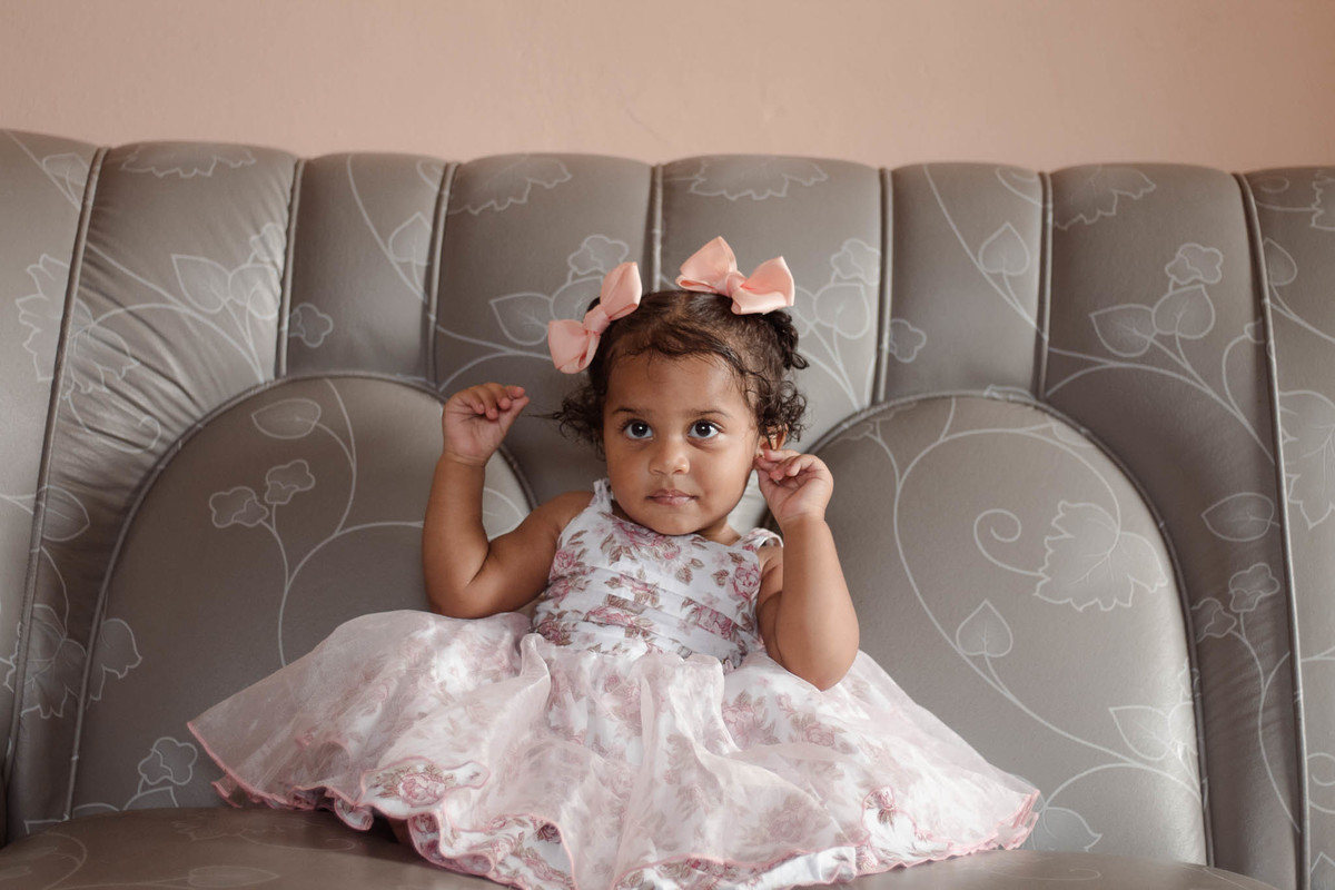 bebê de 1 ano, menina, pele parda, cabelo cacheado castanho e olhos castanhos, vestindo vestido rosa e branco de flores e lacinho rosa na cabeça, sentada em um sofá cinza com a mão esquerda na orelha
