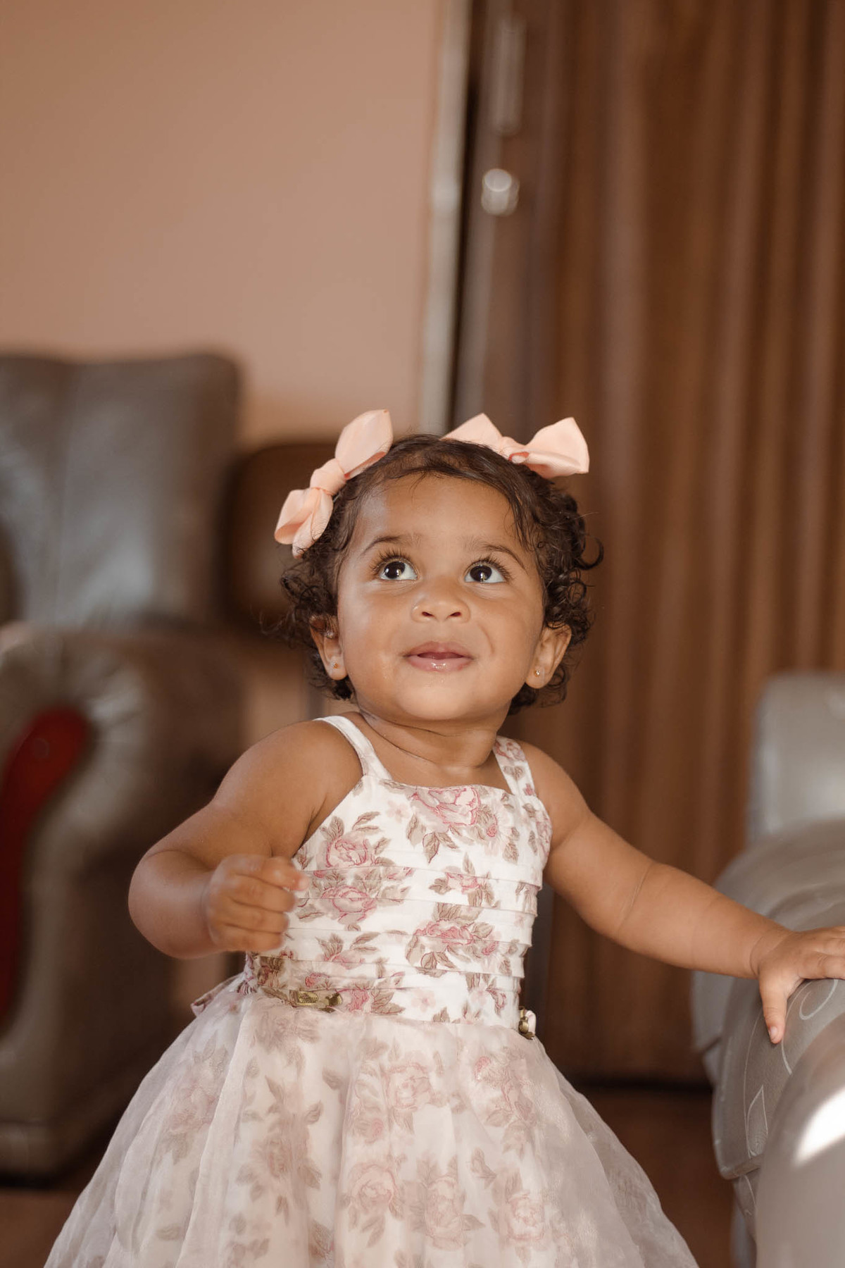 bebê de 1 ano, menina, pele parda, cabelo cacheado castanho e olhos castanhos, vestindo vestido rosa e branco de flores e lacinho rosa na cabeça, olhando para cima
