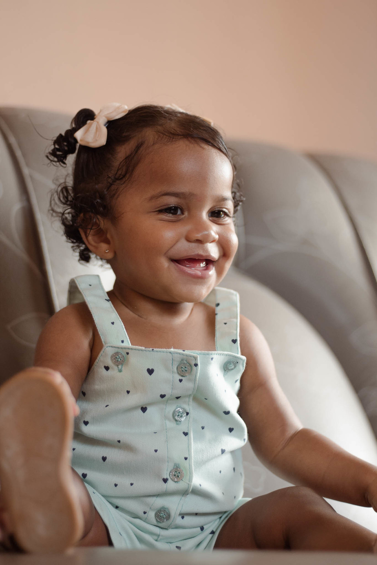 bebê de 1 ano, menina, pele parda, cabelo cacheado castanho e olhos castanhos, vestindo macacão azul e lacinho rosa na cabeça, sorrindo