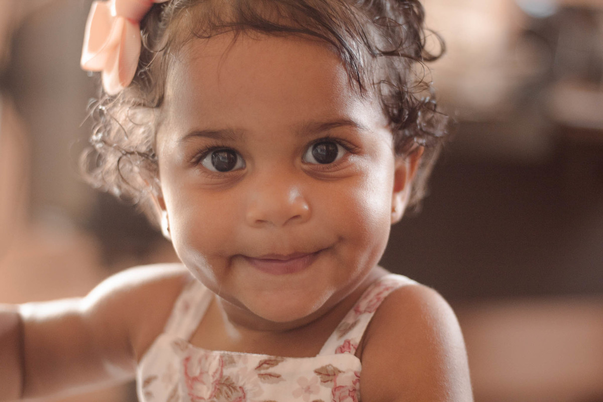 bebê de 1 ano, menina, pele parda, cabelo cacheado castanho e olhos castanhos, vestindo vestido rosa e branco de flores e lacinho rosa na cabeça, sorrindo olhando para a câmera