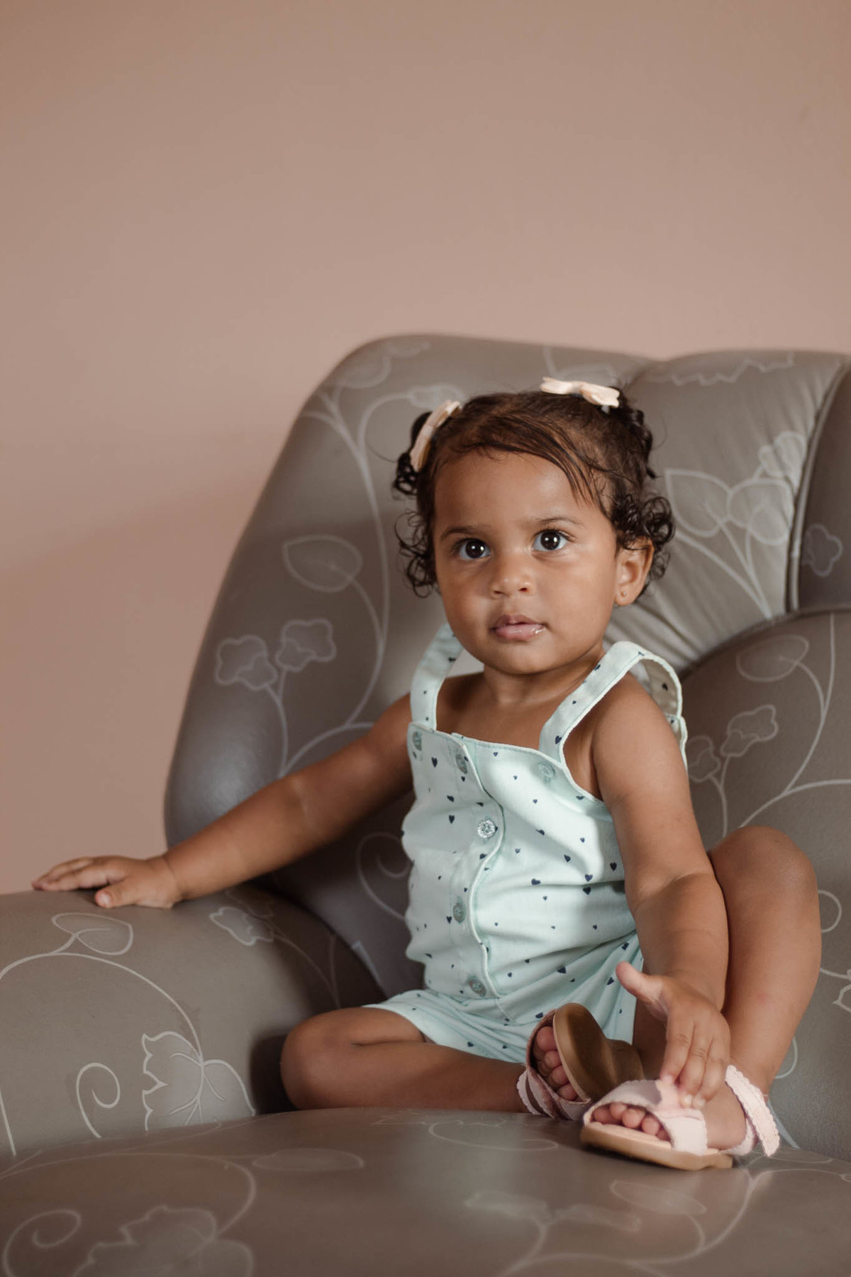 bebê de 1 ano, menina, pele parda, cabelo cacheado castanho e olhos castanhos, vestindo macacão azul e lacinho rosa na cabeça, séria olhando para frente