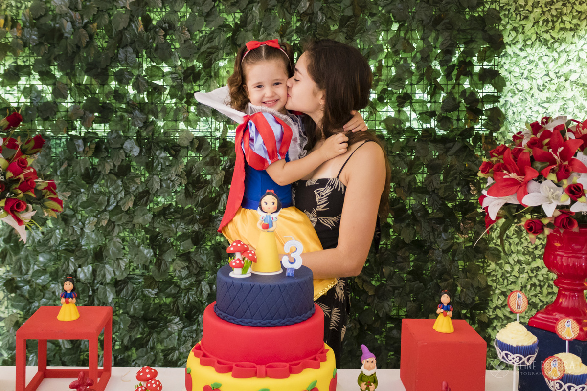 menina vestida de branca de neve ganhando um beijo da irmã na mesa do bolo decorada com o tema Branca de neve e os 7 anões