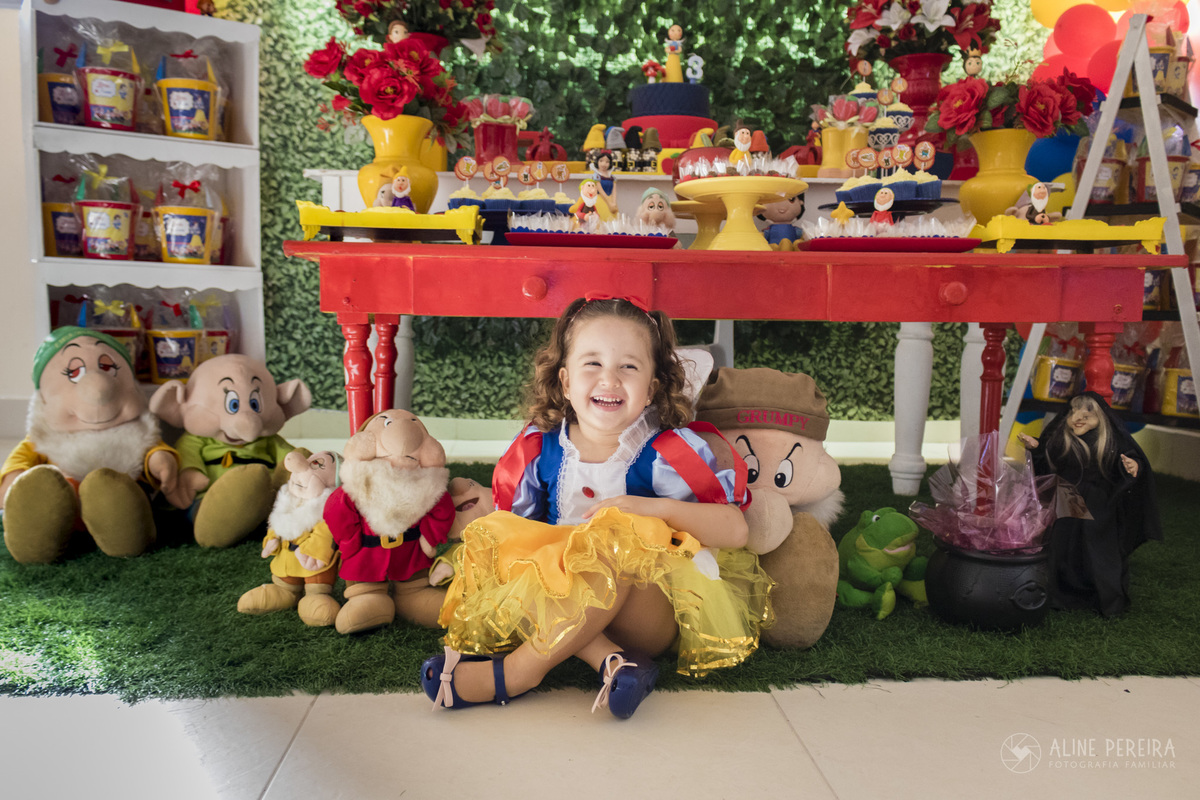 menina vestida de branca de neve ao lado dos 7 anões de pelucia e em frente a mesa do bolo decorada com o tema Branca de neve e os 7 anões