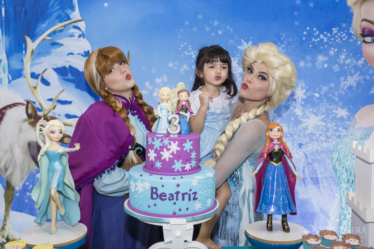 Personagens vivo das princesas Elsa e Ana da Frozen mandando beijinho junto com a aniversariante na mesa do bolo decorada com o tema frozen