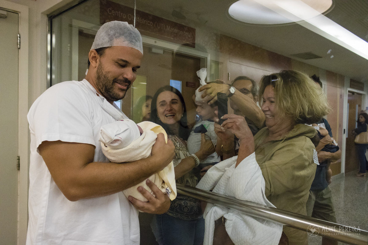 pai emocionado mostrando a filha de dentro do berçário para a família do lado de fora