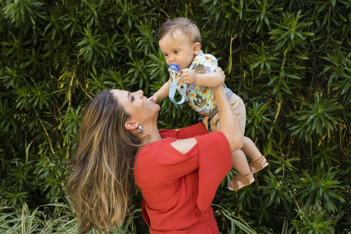 mãe de vestido vermelho levantando o filho para o alto num lugar com muitas plantas