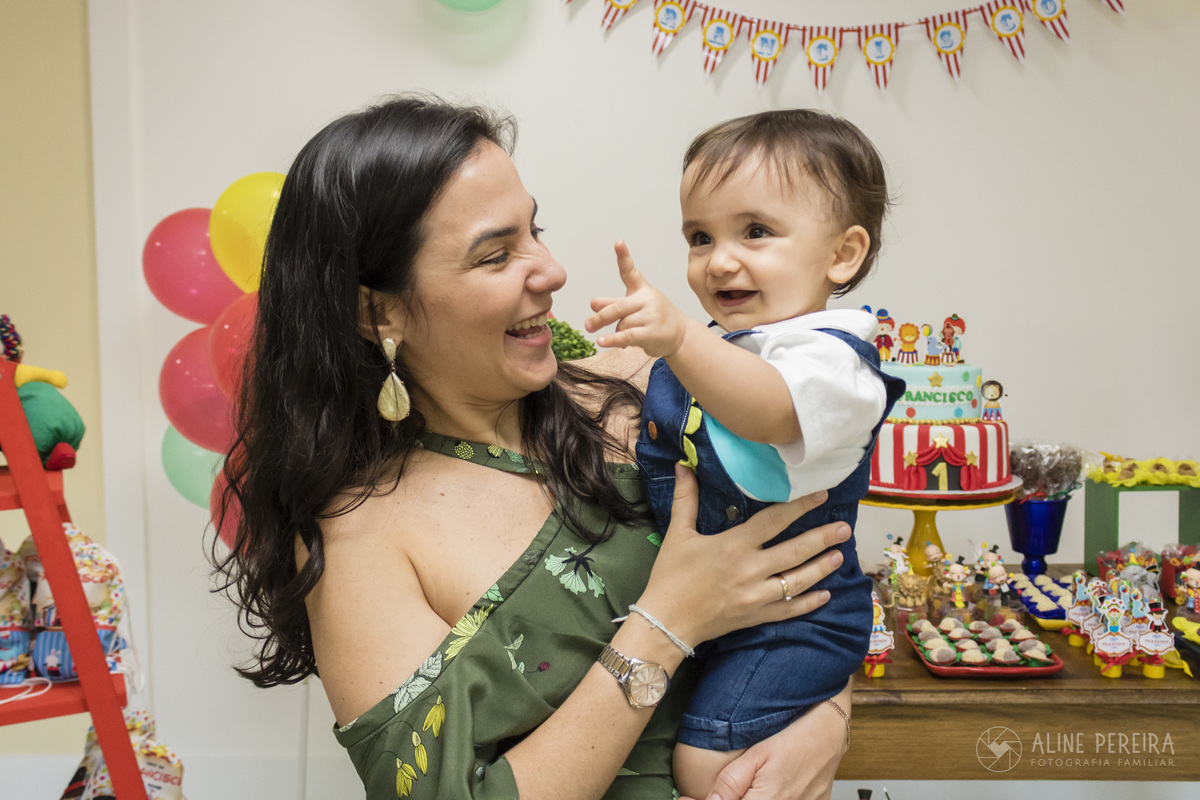 menino no colo da mãe fazendo o numero um com o dedo em frente a mesa do bolo 