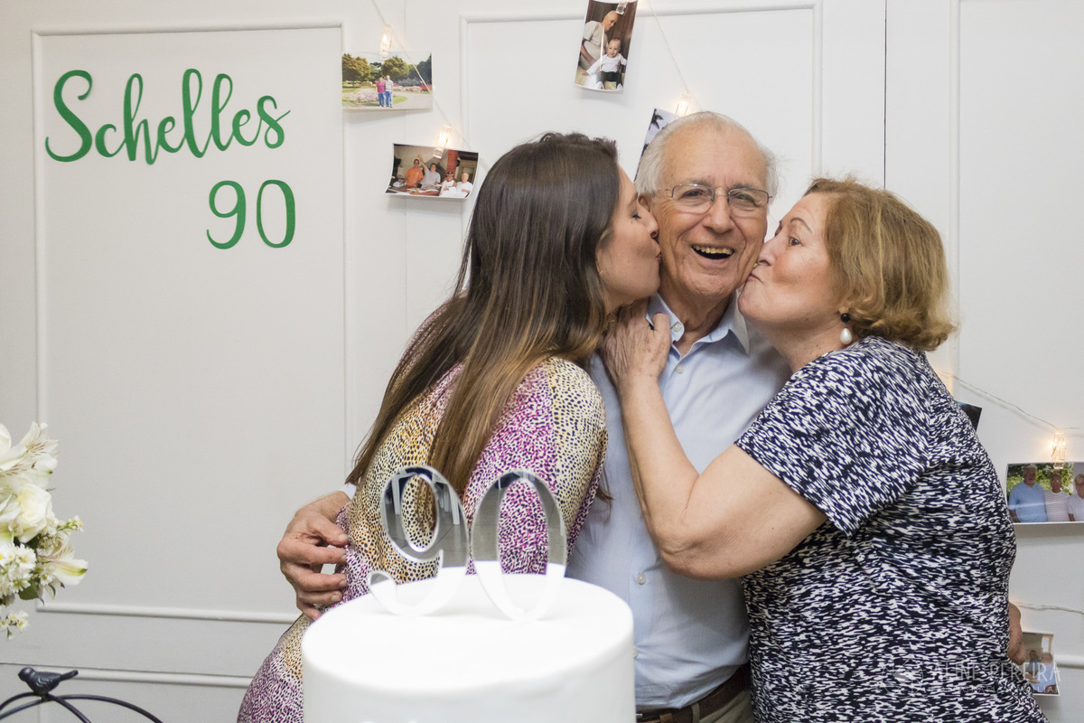 idoso sendo beijado pela mulher e filha na mesa de aniversário dos seus 90 anos