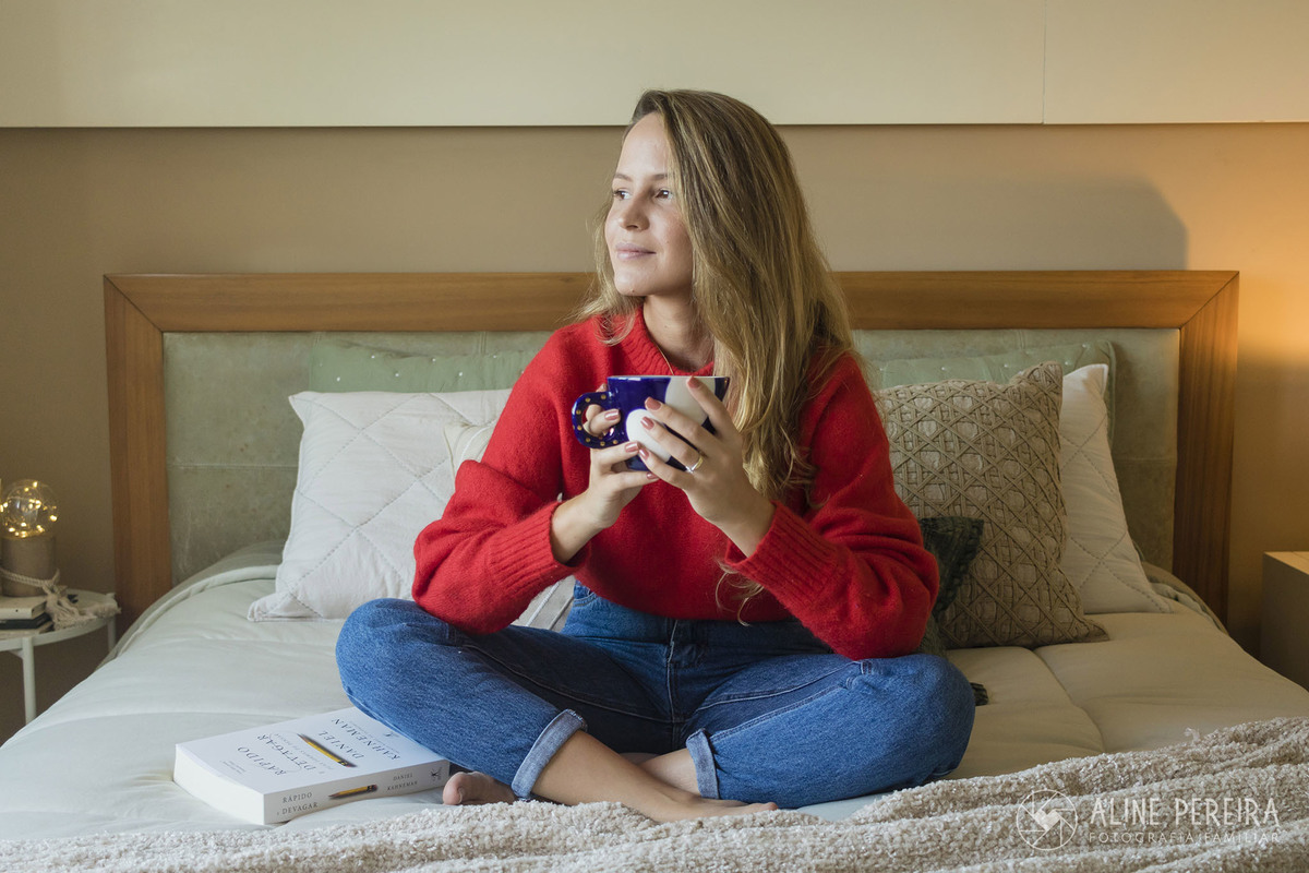 mulher sentada na cama com uma xicara na mão e um livro na outra