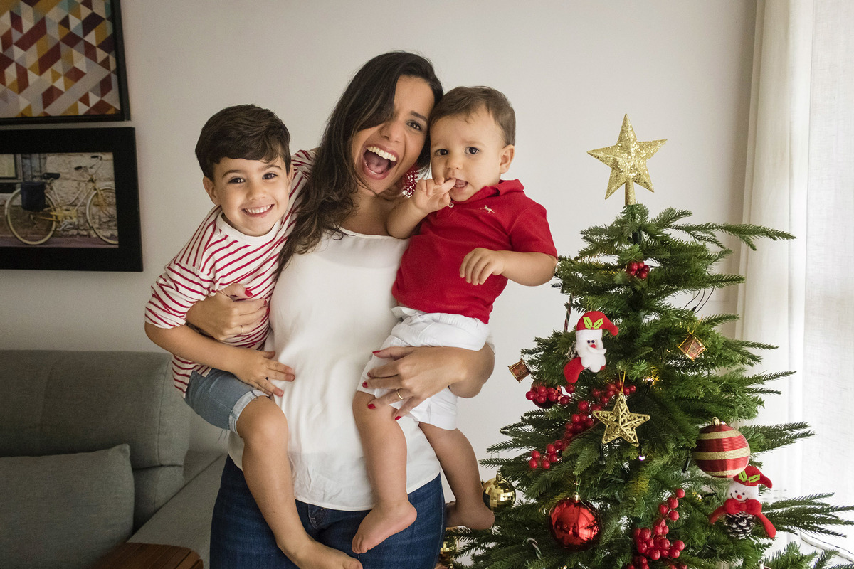 mãe com dois filhos nos braços em frente a árvore de natal