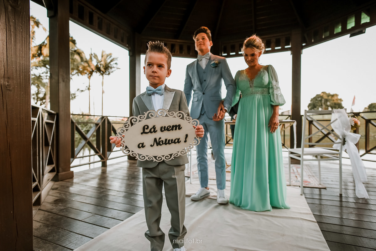 pajem com a placa de lá vem a noiva, e noivo ao fundo emocionado 