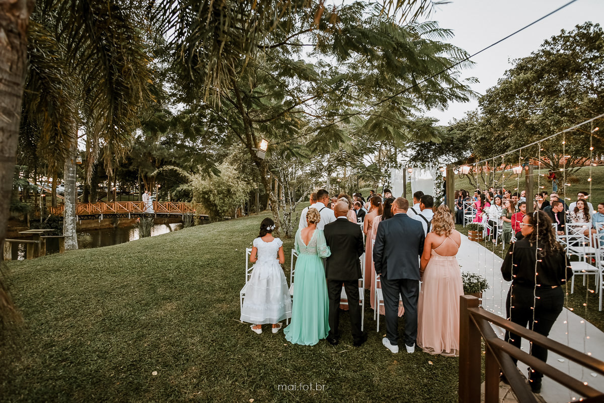convidados de pé, olhando a chegada da noiva na cerimonia, noiva atravessando ponte sobre o lago para chegar na cerimonia de casamento final de tarde de 23/04/2022