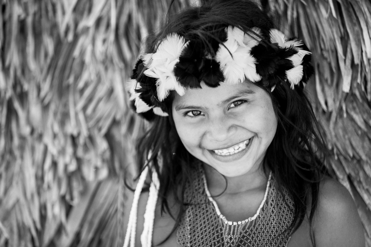 menina índia com arco de penas e flores sorrindo para a câmera, na área indígena do oeste do mato grosso na aldeia parecis,  fotografia por alê carnieri