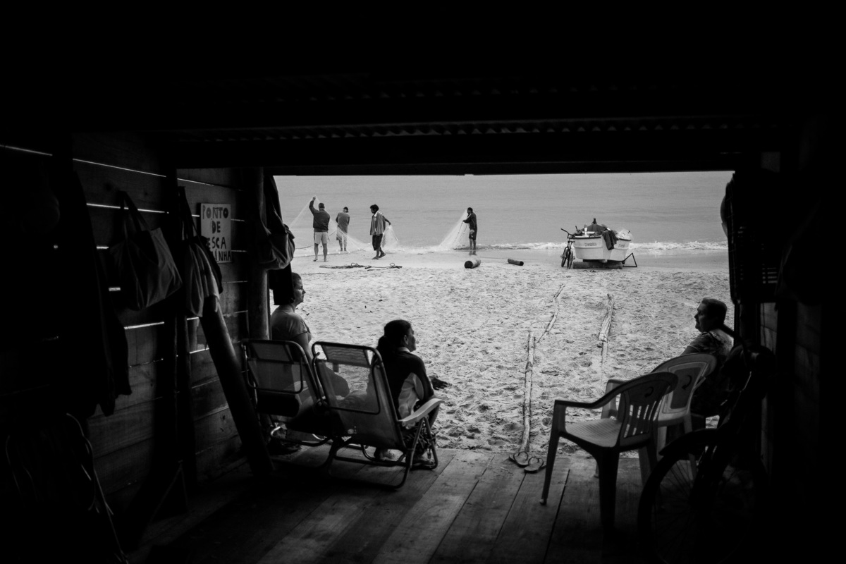 homens puxando rede com peixes, pessoas dentro de cabana de pesca esperando a retirada do barco na pesca da tainha em Florianópolis - SC, fotografia por Alê Carnieri
