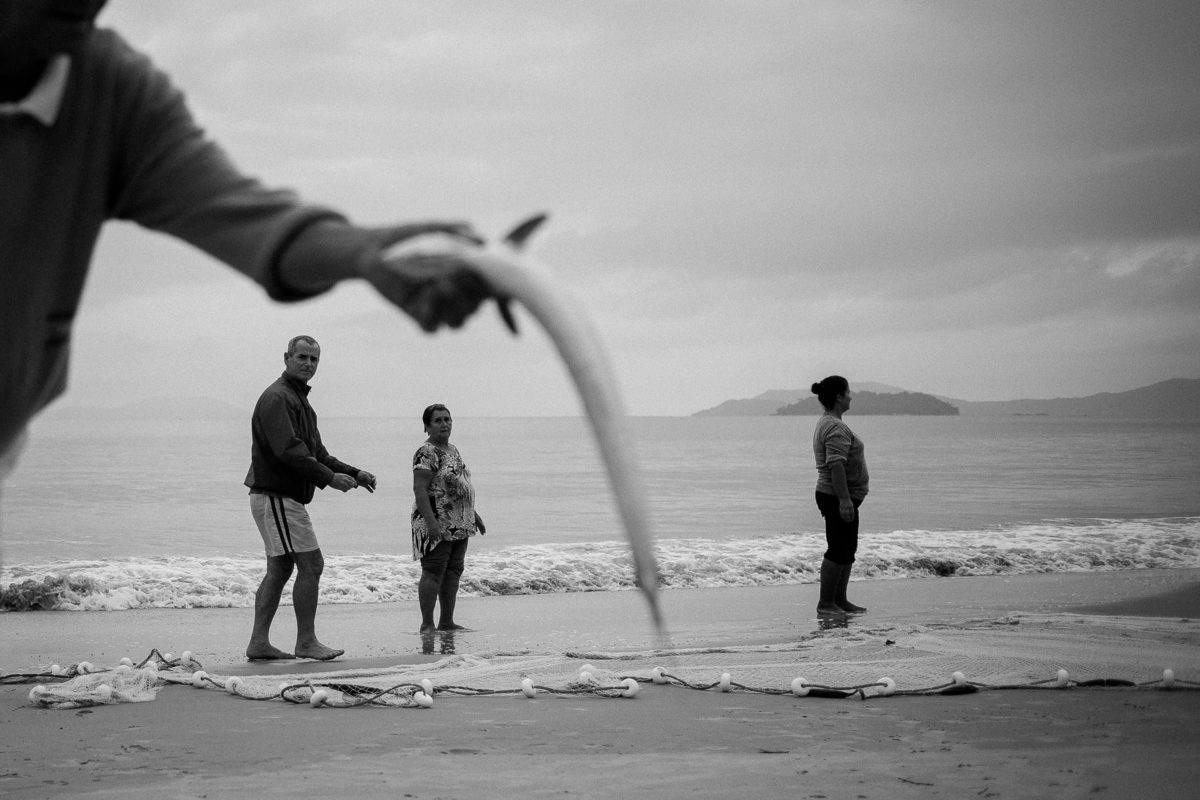 três pessoas na beira do mar, um homem segurando peixe na pesca da tainha em Florianópolis - SC, fotografia por Alê Carnieri