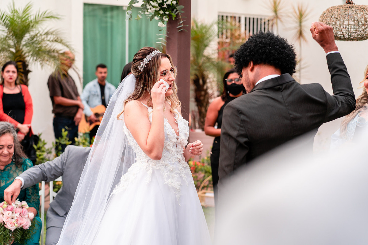 A noiva e o noivo estão de pé de frente um para o outro, durante a cerimônia. A noiva está chorando e o noivo está com a mão fechada, com o punho para cima, de alegria.