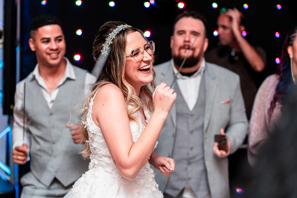 A noiva está rindo e dançando na pista de dança, segurando as mãos próximas ao rosto. Ela usa um vestido de noiva branco e óculos. O fundo é de convidados e luzes coloridas.