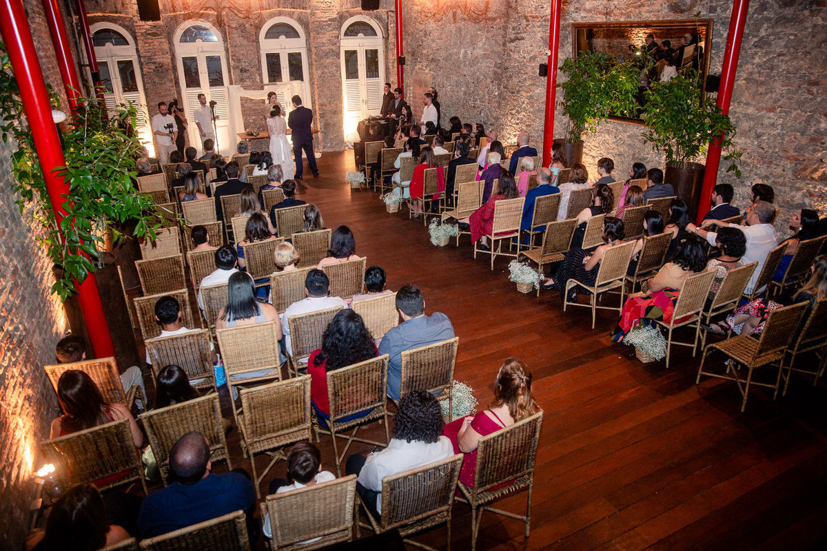 A celebração do casamento vista de cima, com os convidados sentados em cadeiras de vime em um espaço rústico.