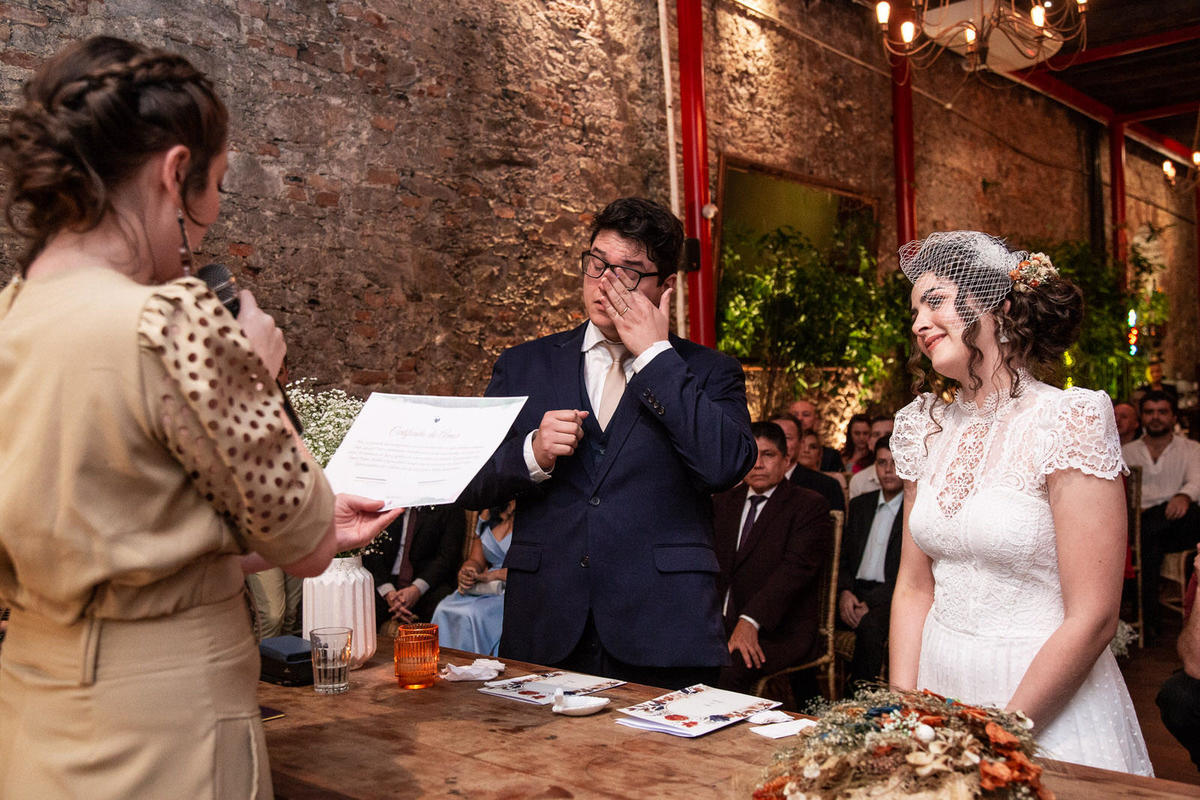 O noivo, em seu terno azul, se emociona e esfrega o rosto com a mão, enquanto a noiva sorri feliz ao seu lado.