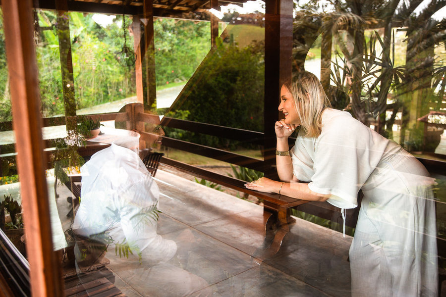 Noiva, usando vestido longo e branco, se apoia em uma bancada de madeira, pensativa e sorridente, em uma varanda com vista para a serra carioca.