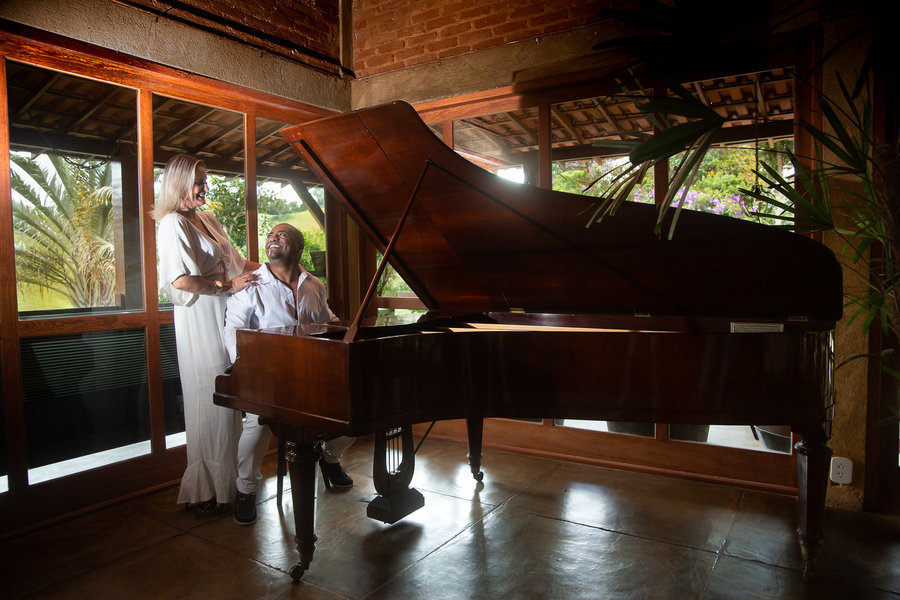 Vista ampla do casal junto a um piano de cauda de madeira, com grandes janelas ao fundo que mostram a vista para a paisagem da serra.