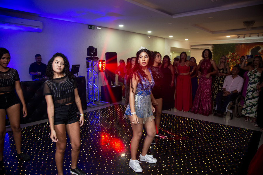 A debutante, em seu segundo vestido, e o grupo de dança fazem uma pose final na pista, com luzes coloridas e o público ao fundo.