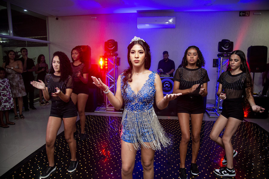 A debutante faz um gesto com as mãos, liderando o grupo de dança na pista, sob a luz dos refletores coloridos.