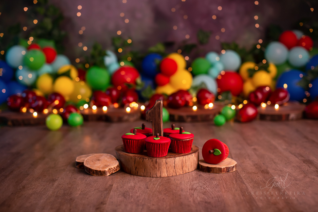 smash the cake branca de neve cupcake formato de maca sobre fatia de madeira a frente de um  fundo temos baloes plantas artificiais e pisca-pisca