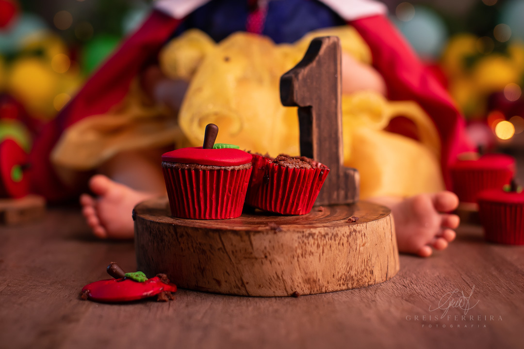 smash the cake branca de neve cupcake formato de maca sobre fatia de madeira a frente de uma bebe de 1 ano com fantasia da branca de neve em um  fundo com baloes plantas artificiais e pisca-pisca