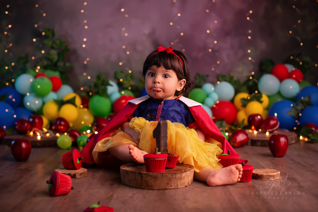smash the cake branca de neve cupcake no formato de maca destruidos sobre fatia de madeira a frente de uma bebe de 1 ano com fantasia da branca de neve em um  fundo com baloes plantas artificiais e pisca-pisca